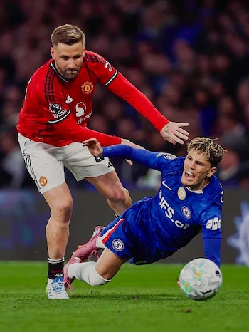 Dos futbolistas en acción; uno con camiseta roja del Manchester United bloquea a otro con camiseta azul del Chelsea que cae al césped junto al balón