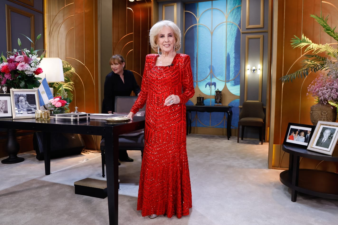 Mirtha Legrand posa sonriente en el set de su programa, luciendo un elegante vestido rojo de lentejuelas junto a un escritorio adornado.
