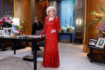Mirtha Legrand, mujer mayor de cabello rubio platinado, sonríe en un set de televisión, vistiendo un largo vestido rojo brillante. Detrás hay un escritorio y una mujer