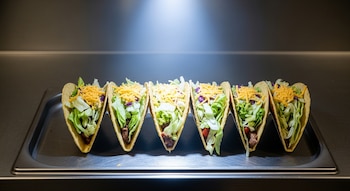 Seis tacos de tortilla crujiente rellenos de carne, lechuga picada y queso rallado, alineados en una bandeja de acero inoxidable bajo un haz de luz fría.