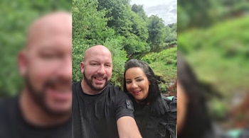 Primer plano de un hombre y una mujer sonriendo, con un río, árboles y vegetación borrosa de fondo.