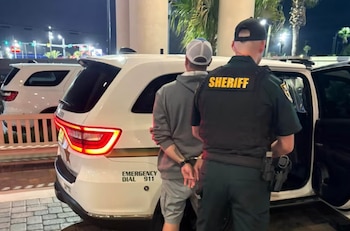 Un hombre esposado es acompañado por un oficial de sheriff, vestido con uniforme y chaleco, junto a un vehículo policial de noche