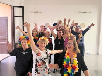 Grupo de personas adultas sonriendo y levantando los brazos en una sala luminosa. Muchos usan collares y diademas de flores coloridas