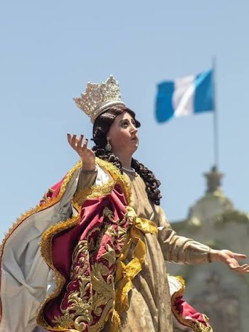 Estatua de una figura femenina coronada con túnica dorada, capa roja y dorada, y manto blanco. Tiene trenzas oscuras y las manos abiertas. Bandera de Guatemala al fondo