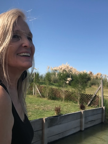 Mujer rubia sonriente de perfil junto a una estructura de madera, con césped y plantas bajo un cielo despejado
