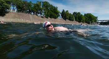 Pessoas usando toucas e óculos de natação em águas abertas, com outros nadadores, muro de pedra, árvores e ponte ao fundo sob o céu azul