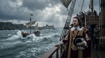 Un hombre con traje del siglo diecisiete está en la cubierta de un barco de madera frente a un mar agitado, otra embarcación y un castillo.