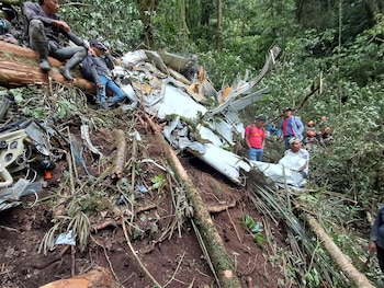 Restos de aeronave blanca entre densa vegetación selvática, ramas y troncos. Varias personas están de pie y sentadas alrededor del lugar