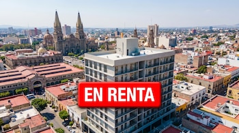 Vista aérea diurna de Guadalajara con un edificio moderno de departamentos y un letrero rojo digital 'EN RENTA'. Al fondo, la Catedral y el skyline urbano.
