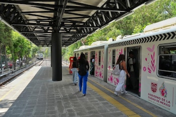 Varias personas caminan hacia y abordan un tren blanco decorado con diseños florales rosas y el logo del Gobierno de la Ciudad de México en una estación al aire libre