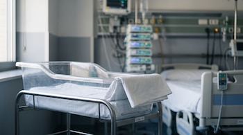 Una cuna de acrílico transparente con una sábana blanca se encuentra junto a una cama de hospital y monitores de control.