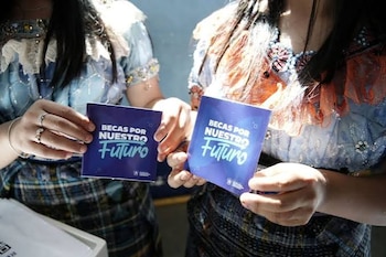 Dos personas sostienen sendas tarjetas moradas con el texto "Becas por Nuestro Futuro" en letras blancas. Una persona usa anillos y pulsera