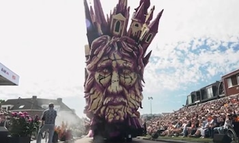 Una enorme carroza con forma de rostro y torres en la parte superior avanza por una calle junto a espectadores sentados en gradas