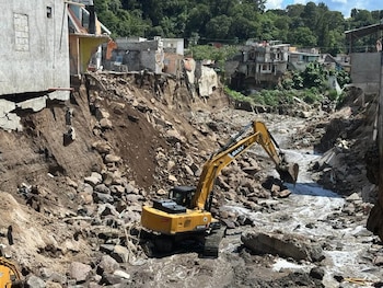 Un río de agua marrón fluye entre dos laderas erosionadas donde hay casas de lámina y concreto construidas cerca del precipicio