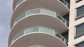 Primer plano de un edificio. Muestra una serie de balcones curvos con barandillas de vidrio y ventanas rectangulares en una pared clara