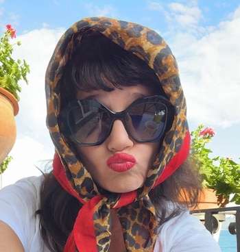 Griselda Siciliani con pañuelo de leopardo y borde rojo, gafas de sol negras, y labios rojos fruncidos. Fondo con plantas, cielo azul y mar