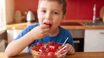 Primer plano de un niño en camiseta azul comiendo una gomita roja de un tazón de dulces de colores sobre una mesa de madera. Fondo de cocina roja.
