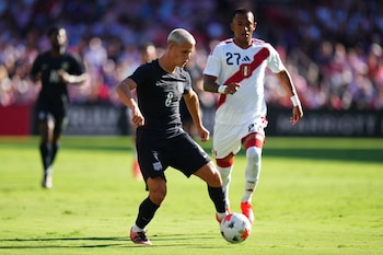 Un jugador de fútbol con uniforme oscuro conduce el balón mientras otro con camiseta blanca y franja roja corre detrás en la cancha