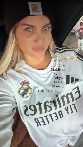Wanda Nara, de cerca, dentro de un coche, lleva un gorro negro y una camiseta blanca del Real Madrid. Su cabello rubio cae sobre sus hombros