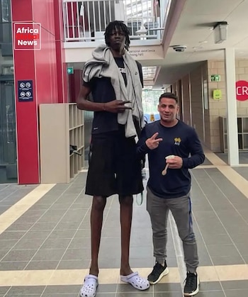 Hombre de piel oscura con dreadlocks, camiseta y bermuda negras, suéter gris sobre hombros y crocs morados. Hombre de piel clara con camiseta azul, pantalón gris y zapatillas negras. Ambos hacen el signo de paz en un interior