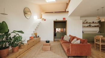 Una sala de estar luminosa con paredes blancas y un techo inclinado. Se ve una escalera flotante blanca, un sofá naranja, plantas y una cocina de gabinetes verdes.