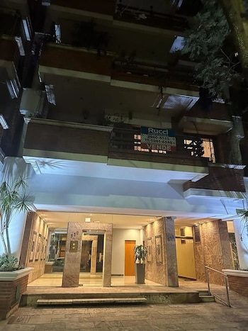 Fachada nocturna de un edificio de apartamentos con entrada iluminada y dos plantas superiores, mostrando un cartel de 'Vende' en un balcón