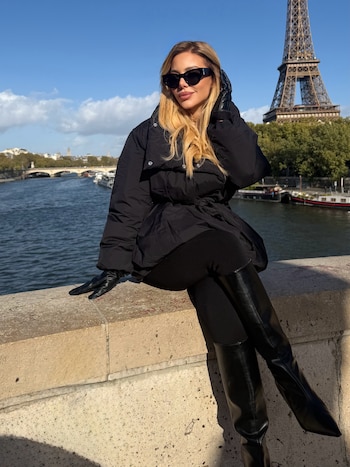 Una mujer vestida de negro con gafas de sol posa sentada sobre un muro de piedra frente al río Sena y la Torre Eiffel