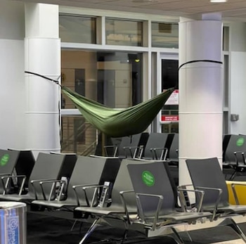 Hamaca verde colgada entre dos pilares blancos. Filas de asientos negros, bote de basura y ventanas al fondo en un aeropuerto