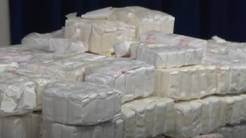 Primer plano de una gran pila de paquetes rectangulares envueltos en material blanco o claro, algunos con pequeñas marcas rojas, sobre una superficie