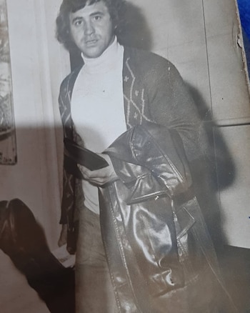Hombre de cabello oscuro con suéter de cuello alto y cárdigan estampado, sostiene chaqueta de cuero y un objeto rectangular. Foto en blanco y negro