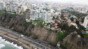Vista aérea de acantilados de tierra con edificios en la parte superior, una autopista con autos en la base y olas del océano