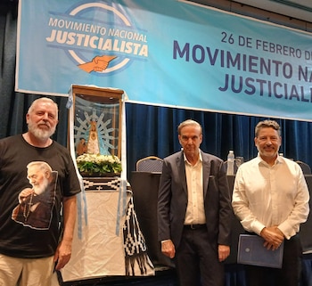 Tres hombres, Esteban Castro, Miguel Pichetto y Guillermo Moreno, posan de pie frente a un banner azul y una imagen religiosa en una urna de vidrio