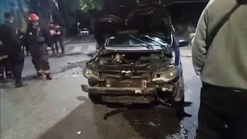 Vehículo oscuro chocado con capó abierto y parachoques roto en una calle oscura, con líquidos y escombros. Personal de emergencia y policías alrededor