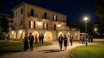 Villa Mussolini, edificio blanco iluminado, en la noche. Personas vestidas de gala caminan por senderos de grava hacia la entrada, con farolas encendidas.
