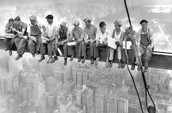 Fotografía en blanco y negro de once hombres sentados en hilera sobre una viga de acero a gran altura sobre una ciudad