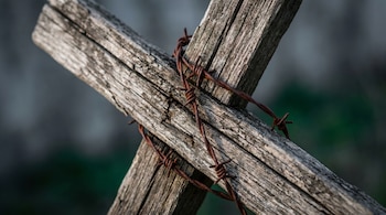 Primer plano de una cruz de madera con textura rugosa y grietas, cruzada diagonalmente por alambre de púas oxidado. El fondo está desenfocado en tonos oscuros.