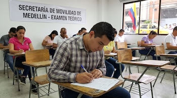 Varias personas sentadas en pupitres escriben en hojas. Un cartel dice 'Secretaría de Movilidad de Medellín - Examen Teórico'. Una bandera de Colombia se ve por la ventana.