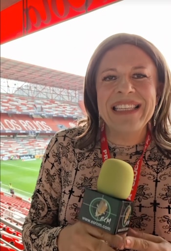 Primer plano de la reportera Roxana Olarte sonriendo, sosteniendo un micrófono, con las gradas rojas de un estadio al fondo