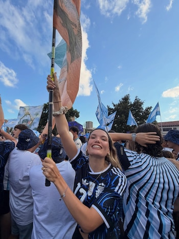 Mujer rubia con camiseta de la Selección Argentina de fútbol sonríe y levanta un asta con una bandera entre una multitud; varias banderas celestes y blancas en el fondo