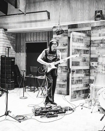 Un hombre con audífonos toca una guitarra eléctrica de pie frente a pedales de efectos, junto a un micrófono y una batería en un estudio
