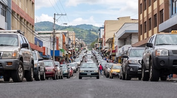 Calle con edificios a ambos lados, muchos coches en el tráfico, personas en las aceras, postes de luz con cables y montañas verdes al fondo bajo nubes.