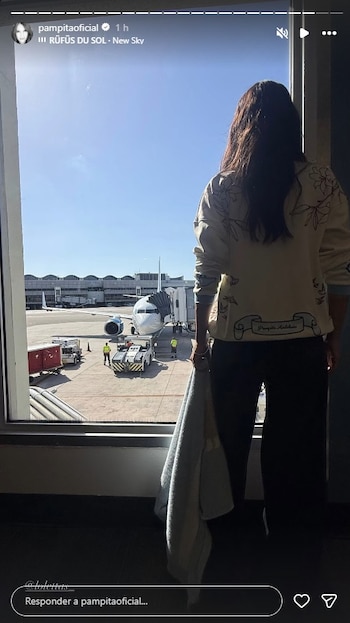 Vista trasera de una persona con cabello oscuro y sudadera clara con dibujos, mirando por la ventana de un aeropuerto a un avión blanco y azul en la pista