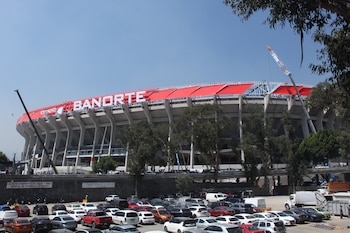 Vista exterior del Estadio Azteca con el logo "BANORTE" y un techo rojo. Se observan grúas de construcción, árboles y un estacionamiento con vehículos