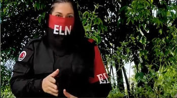 Persona con ojos visibles, rostro cubierto por máscara roja y negra con "ELN", uniforme oscuro con insignia del grupo, frente a densa vegetación verde