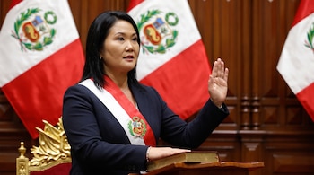 Mujer asiática en primer plano, viste traje oscuro y banda presidencial roja y blanca, mano derecha levantada, mano izquierda sobre libro en podio, banderas peruanas al fondo.