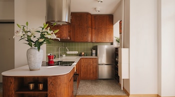 Cocina moderna con gabinetes de madera, encimeras blancas y azulejos verdes. Se ven un refrigerador de acero, lirios blancos en jarrón y una cafetera roja.