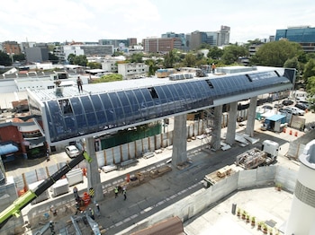 Vista aérea de una estructura elevada de transporte en construcción con trabajadores, grúas, materiales de obra y vehículos en una calle urbana con edificios