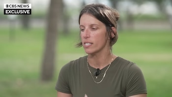 Katie Thomas, una mujer caucásica con cabello castaño recogido y camiseta verde oliva, habla en una entrevista. Un micrófono de solapa está en su camisa. Fondo verde borroso