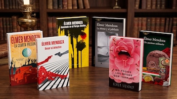 Seis livros com capas e textos coloridos, um abajur antigo, uma mesa de madeira e uma estante ao fundo.