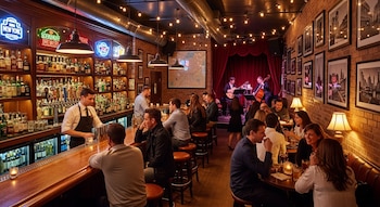 Un bar concurrido con paredes de ladrillo, estanterías de licor, un barman, clientes sentados y de pie, y músicos en un escenario con cortinas rojas.